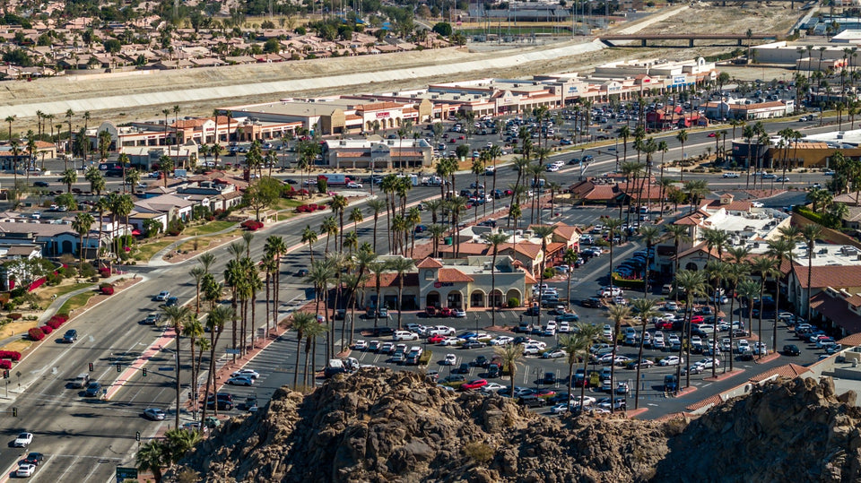 Elevated view of Highway 111 in La Quinta, with shopping centers, palm trees, and the desert ridge in the foreground.