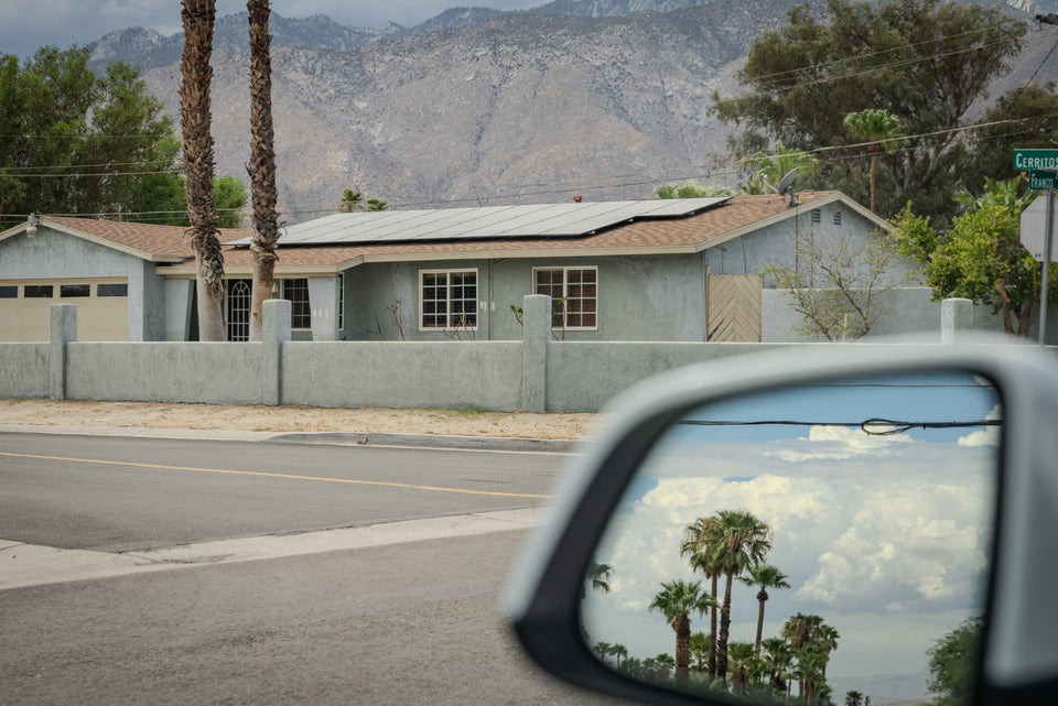 A single-story home with rooftop solar panels in Desert Park Estates, Palm Springs, with palm trees, mountain views, and palms reflected in a car mirror.