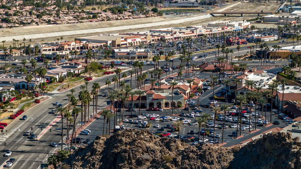Elevated view of Highway 111 in La Quinta, with shopping centers, palm trees, and the desert ridge in the foreground.