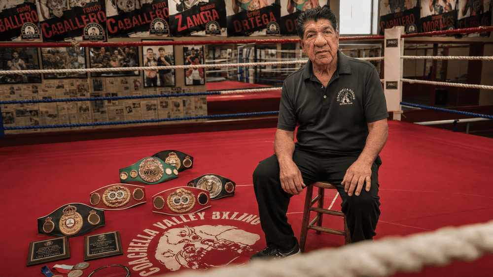 Lee Espinoza seated inside the Coachella Valley Boxing Club ring beside championship belts and the gym’s canvas logo.