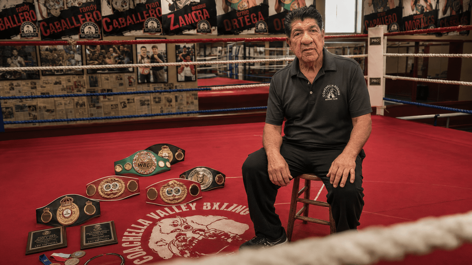 Lee Espinoza seated inside the Coachella Valley Boxing Club ring beside championship belts and the gym’s canvas logo.