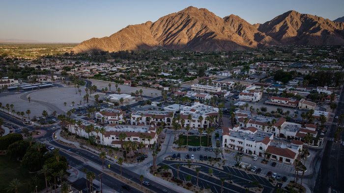 Aerial view of Old Town La Quinta at sundown in late May, showcasing the village-style shopping district, palm-lined streets, and the Santa Rosa Mountains glowing in golden evening light.