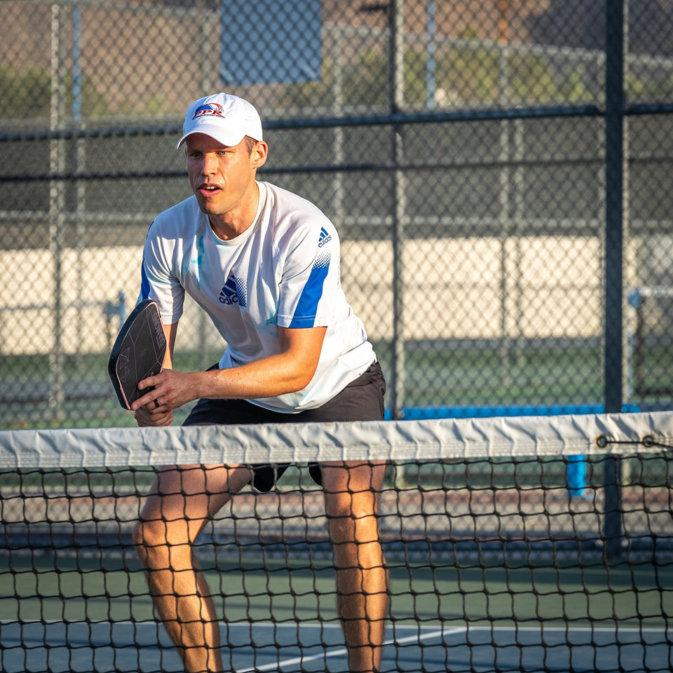 Pickleball player in a white cap preparing for a shot near the net on an outdoor court.