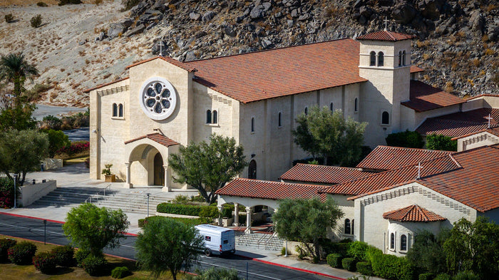 Aerial view of St. Francis of Assisi Catholic Church in La Quinta, California, showing its red tile roof, bell tower, pale exterior walls, and Santa Rosa Mountain backdrop.