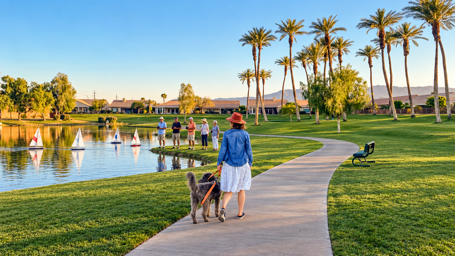 A woman walking a dog beside model sailboats on a Sun City Palm Desert lake; AI-enhanced from Mark Miller’s original photograph.