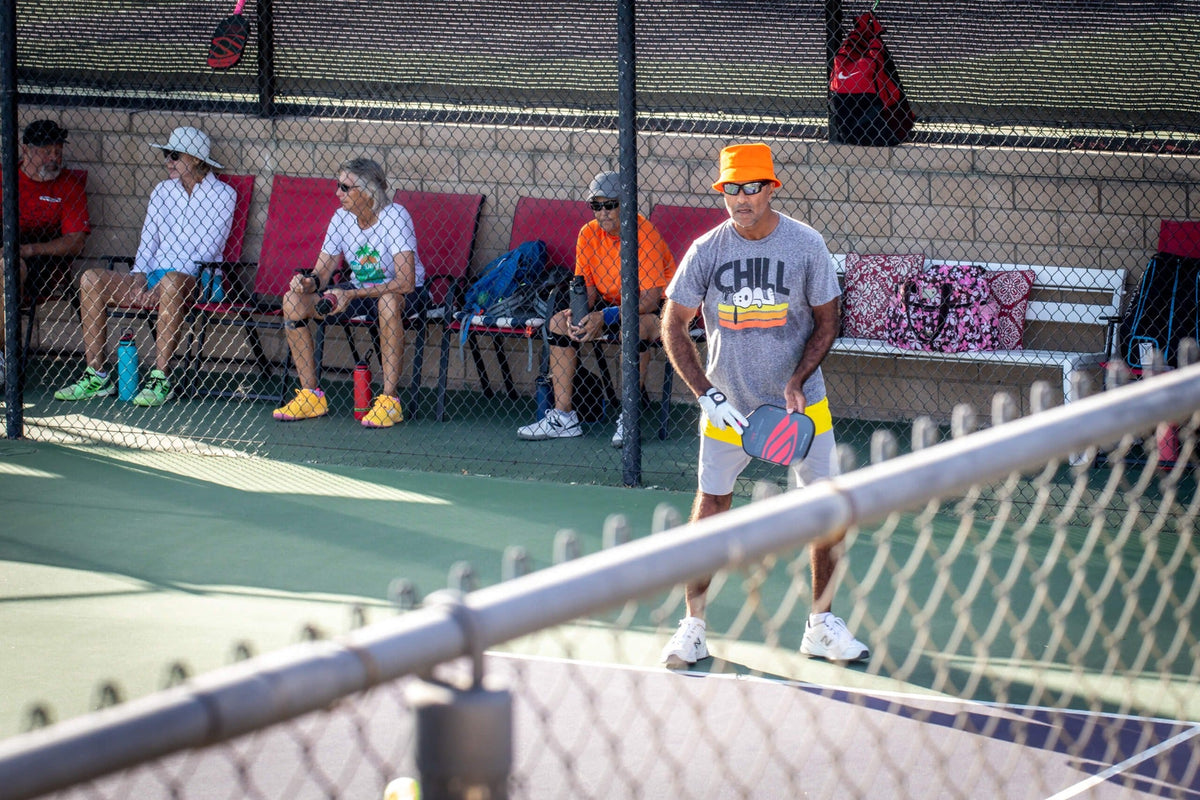 Pickleball player in an orange hat on court at Palm Desert Resort, with spectators seated behind the fence.
