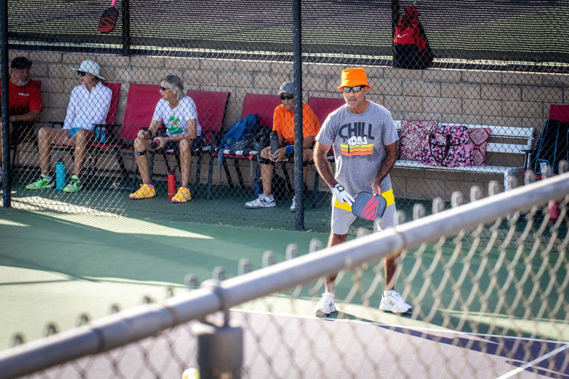 Pickleball player in an orange hat on court at Palm Desert Resort, with spectators seated behind the fence.
