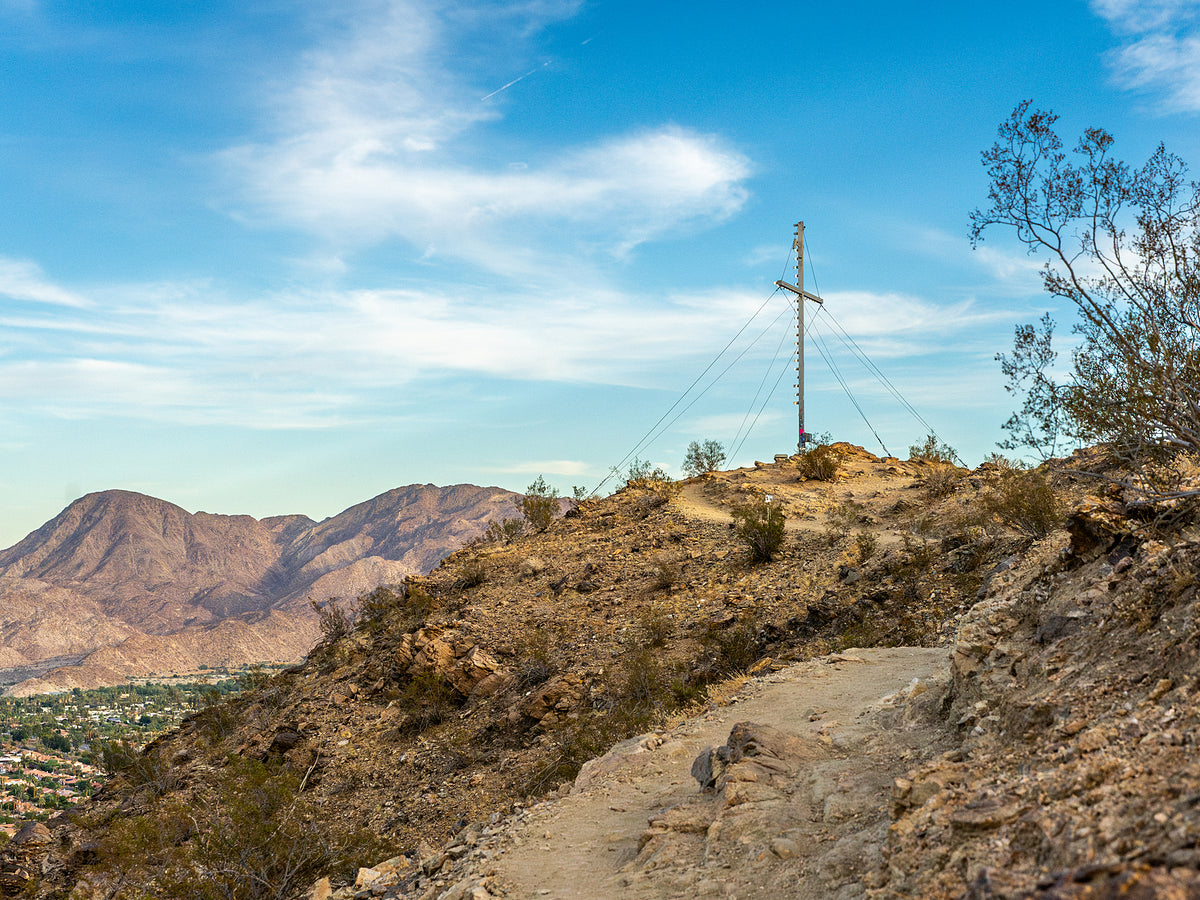 A hiking trail in Palm Desert, CA with a cross on a pole in the background and mountains in the distance.