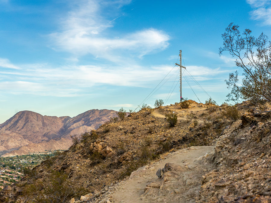 A hiking trail in Palm Desert, CA with a cross on a pole in the background and mountains in the distance.