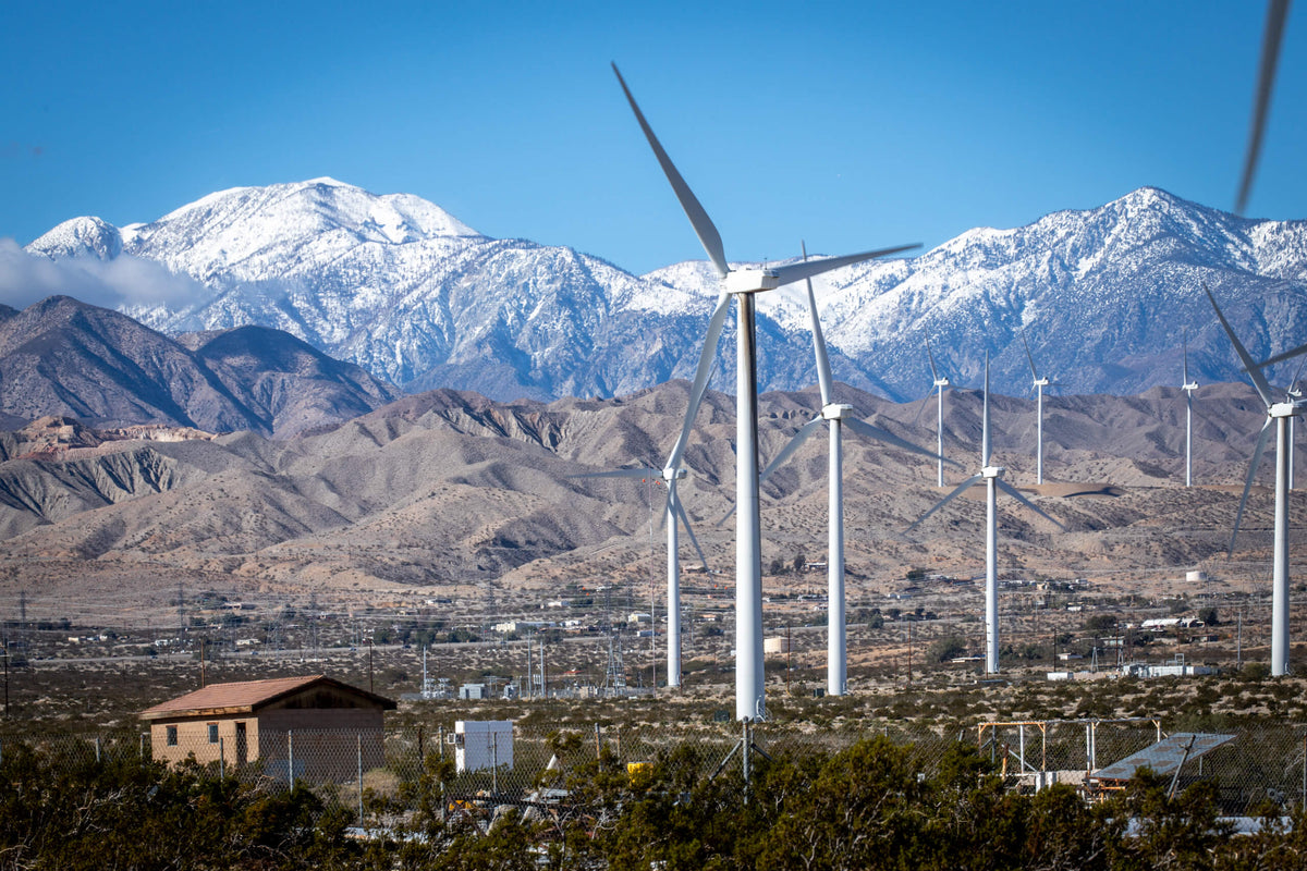 Wind turbines in San Gorgonio Pass with desert hills and snow-covered San Bernardino Mountains beyond.