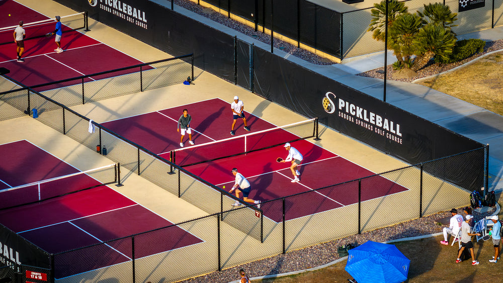 A group of people playing pickleball on a court with red and black markings and a black fence. There is a sports ball visible in the air, and a few players are wearing white tops.