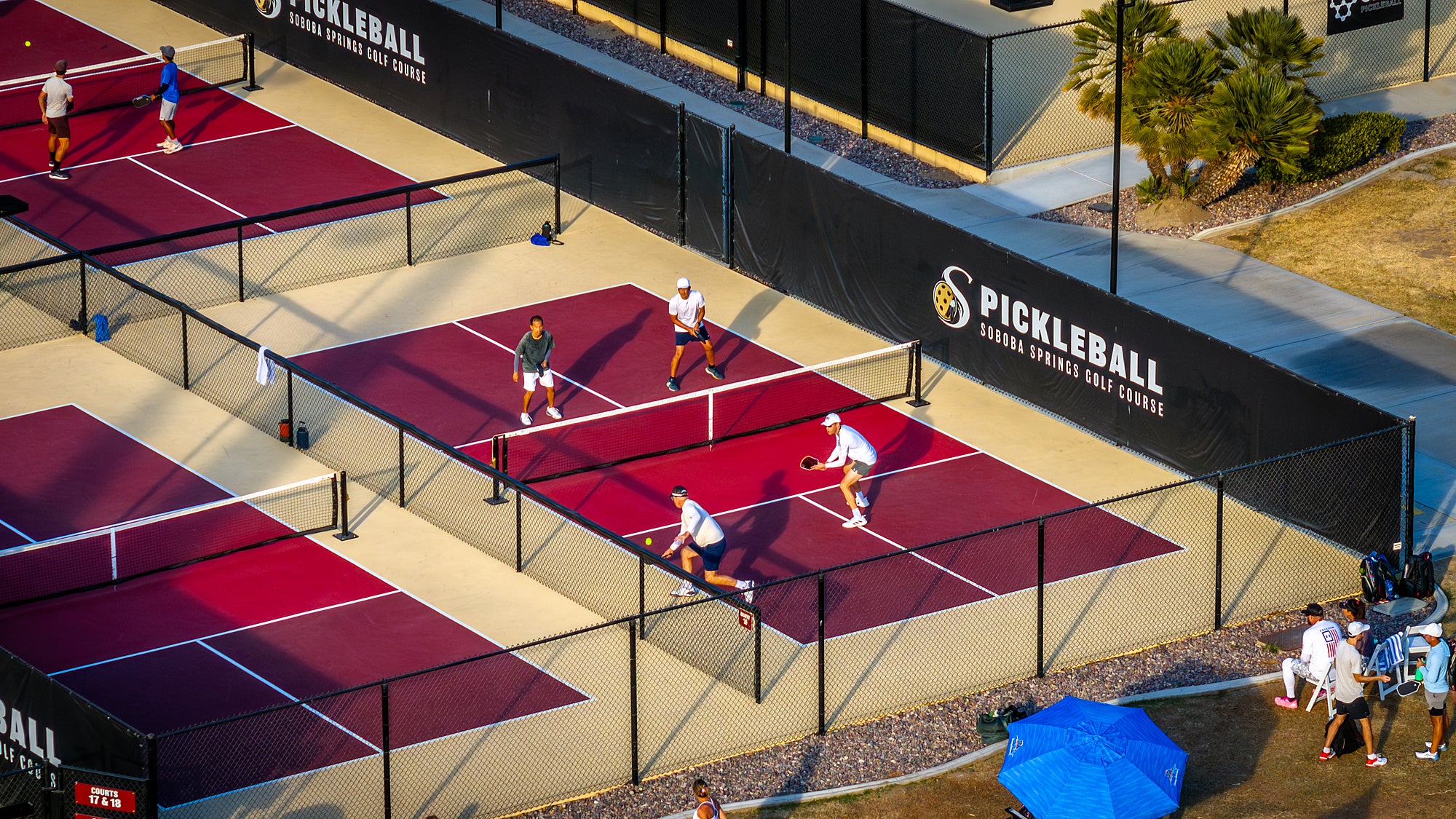 A group of people playing pickleball on a court with red and black markings and a black fence. There is a sports ball visible in the air, and a few players are wearing white tops.