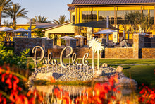 An outdoor view of Trilogy Polo Club featuring a water feature with rocks and plants, leading up to a building with a palm tree and outdoor seating areas.