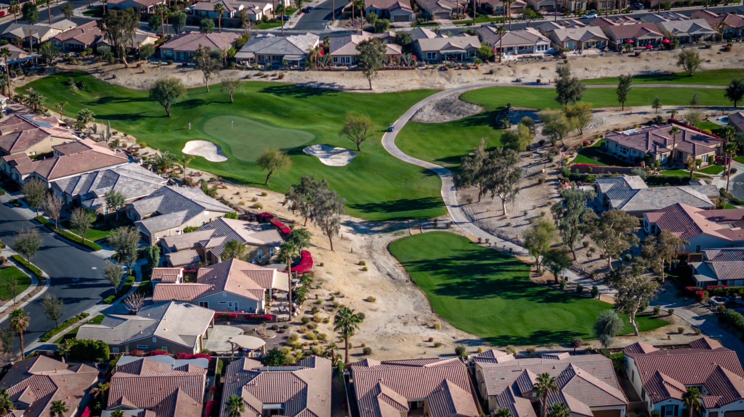 Aerial view of Trilogy La Quinta golf course with green spaces and sand bunkers, surrounded by residential houses, with a clear sky in the background.