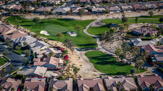 Aerial view of Trilogy La Quinta golf course with green spaces and sand bunkers, surrounded by residential houses, with a clear sky in the background.