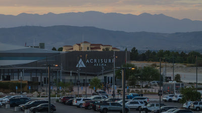 Close-up of Acrisure Arena's exterior sign at sundown, with palm trees, parked cars and mountains in the background.
