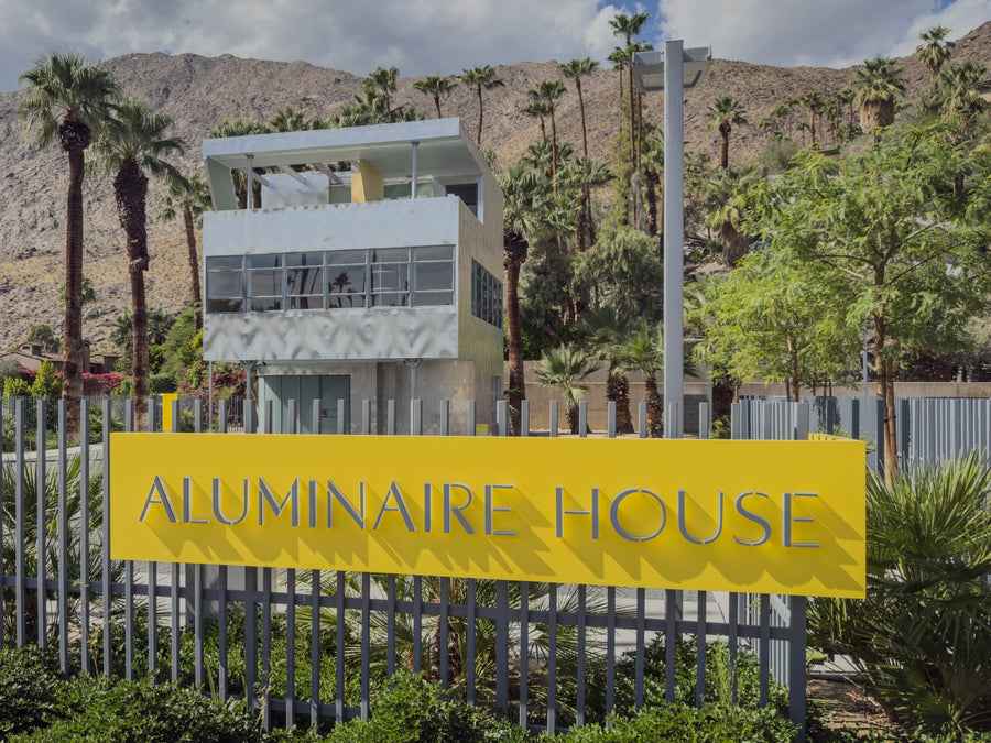 Aluminaire House behind its bright yellow sign in Palm Springs, with palms and mountains beyond. Photograph by Mark Miller, October 20, 2026.