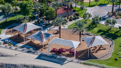 Shade sails cover the playground at Rancho Mirage Community Park beside palm trees and a curving walking path.